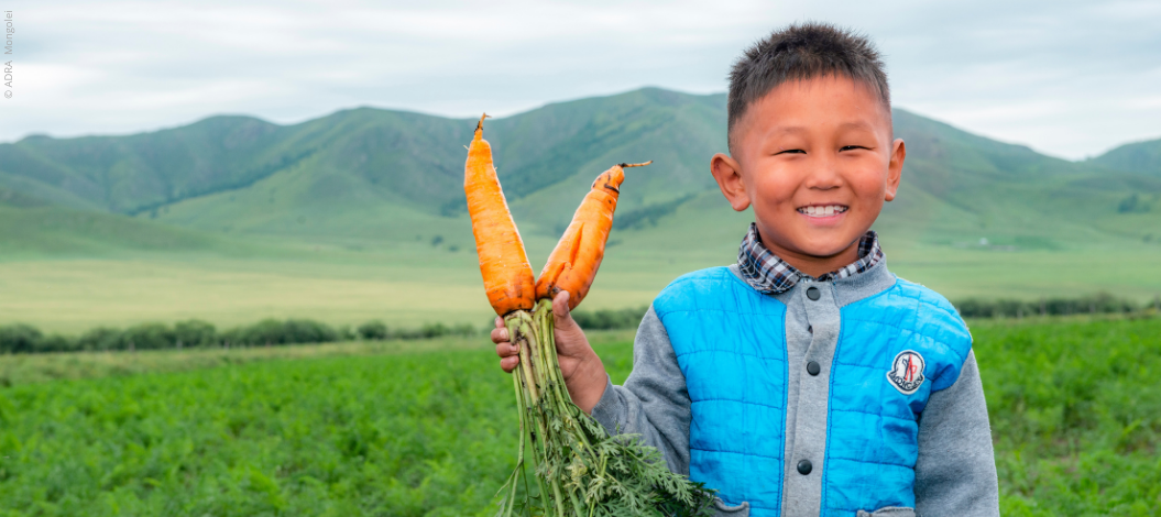 Ein Junge aus der Mongolei steht in einem weiten Feld und hält frisch geerntete Karotten in den Händen, die er stolz in die Kamera zeigt. Im Hintergrund ist die weite Landschaft der Mongolei zu sehen.